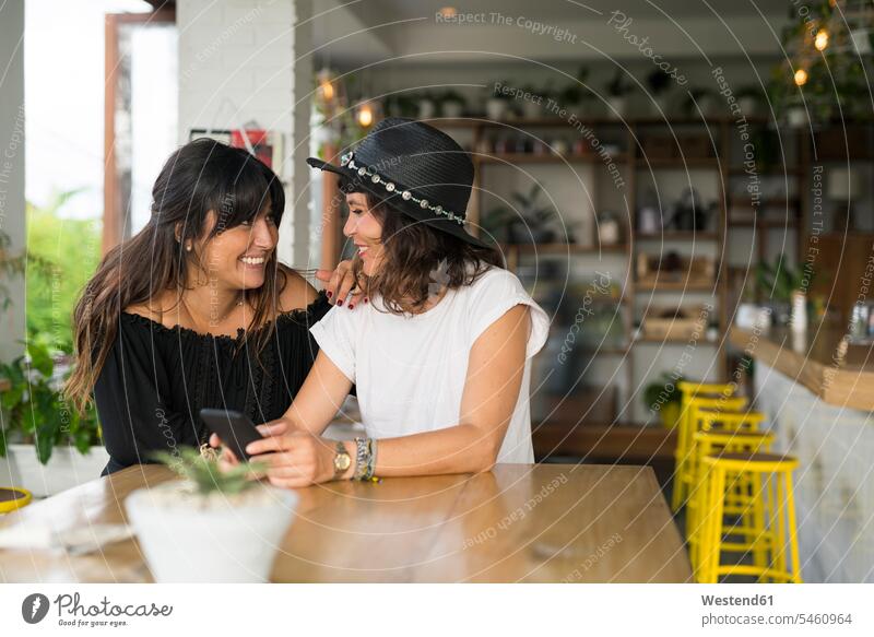 Two female friends smiling at each other in cafe with smartphone woman females women Smartphone iPhone Smartphones smile mate friendship Adults grown-ups