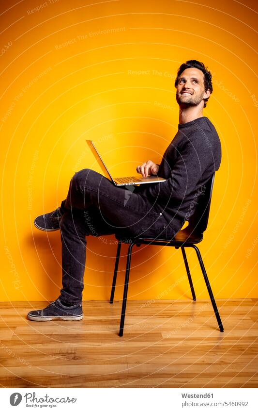 Handsome man looking away while sitting with laptop on chair color image colour image coloured background colored background studio shot studio photograph