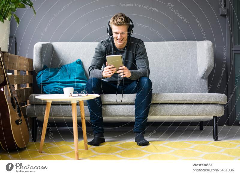 Smiling young man with guitar, tablet and headphones sitting on couch guitars Seated smiling smile men males digitizer Tablet Computer Tablet PC