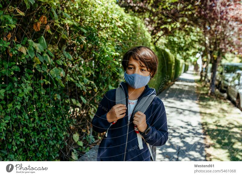 Schoolboy wearing mask looking away while standing by plants on footpath color image colour image Spain casual clothing casual wear leisure wear casual clothes