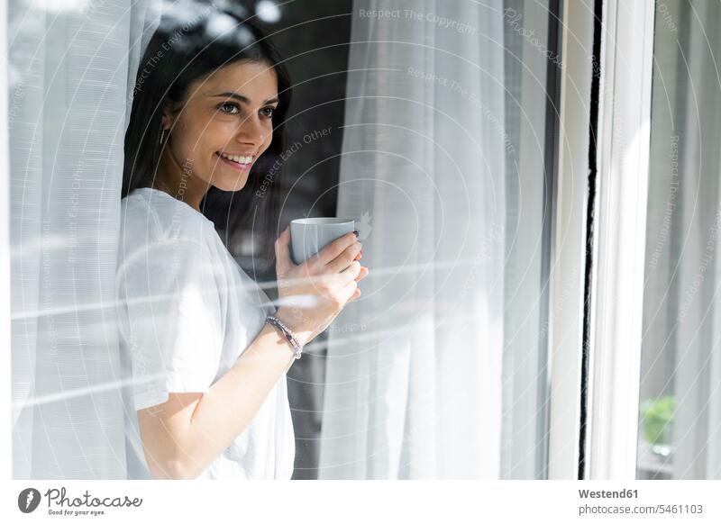 Smiling young woman with cup of coffee behind windowpane human human being human beings humans person persons celibate celibates singles solitary people