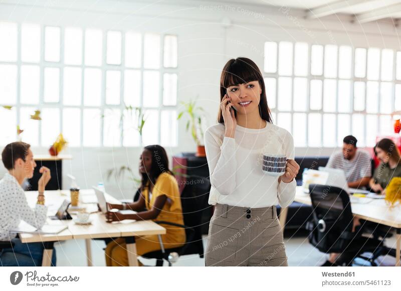 Portrait of smiling young woman on cell phone in office with colleagues in background portrait portraits on the phone call telephoning On The Telephone calling