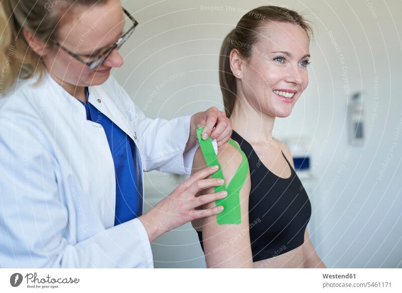Female doctor applying tape to shoulder of patient in medical practice Germany consultation consultations doctor's overall lab coat prevention expertise