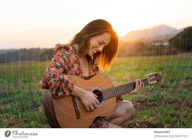 Smiling young woman playing guitar on meadow during sunset color image colour image outdoors location shots outdoor shot outdoor shots sunsets sundown