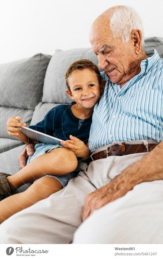 Portrait of happy little boy with digital tablet sitting besides his grandfather on the couch at home portrait portraits happiness grandpas granddads