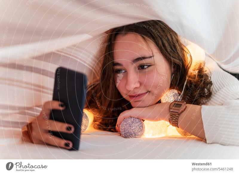 Smiling teenage girl with chain of lights using cell phone underneath bedcover Bed - Furniture beds telecommunication phones telephone telephones cell phones