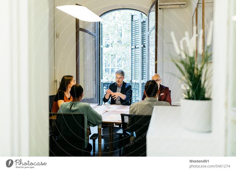 Businessman gesturing while discussing with coworkers at table in office seen through doorway Businessmen Business man Business men businesswear business attire