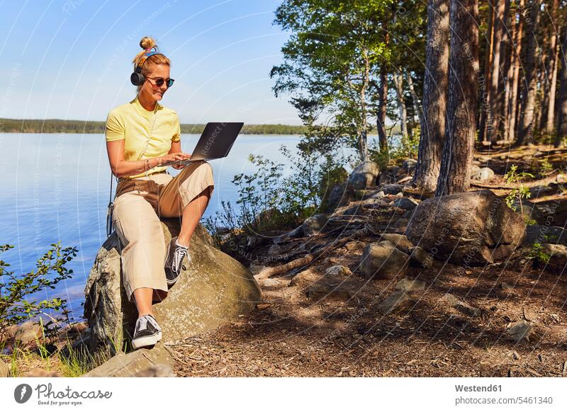 Mid adult woman wearing sunglasses using laptop while sitting against lake in Tiveden National Park, Sweden color image colour image Scandinavia