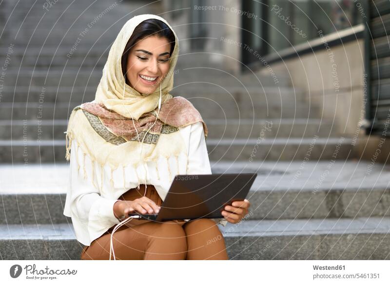 Spain, Granada, young muslim woman wearing hijab using laptop sitting on urban steps headscarf head scarf head scarves Head Scarf head cloths headscarves