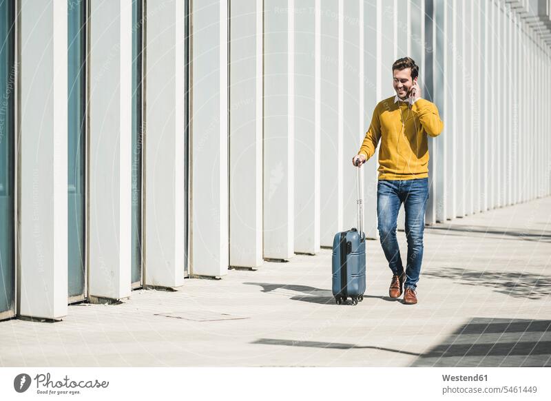 Smiling young man with rolling suitcase and earphones in the city on the go touristic tourists business life business world business person businesspeople