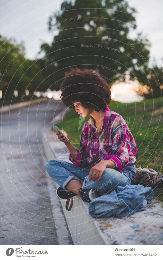 Young woman with afro hairdo sitting on curb using smartphone human human being human beings humans person persons curl curled curls curly hair bags
