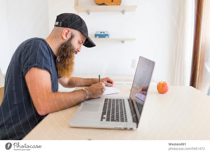 Bearded young man sitting at desk with laptop writing down something desks write beard Seated men males noting Laptop Computers laptops notebook Table Tables