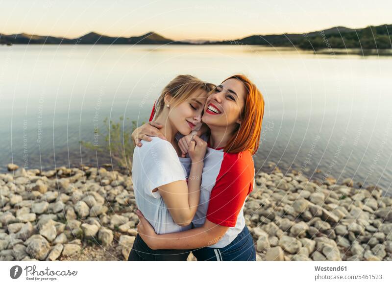 Happy woman embracing girlfriend while standing by lake during sunset color image colour image outdoors location shots outdoor shot outdoor shots sunsets