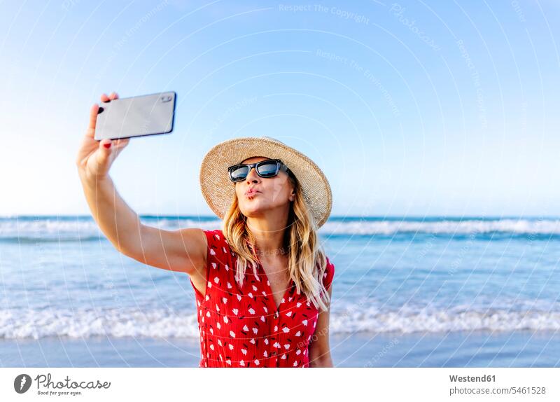 Blond woman wearing red dress and hat and using smartphone and taking a selfie at the beach, making a kissing mouth telecommunication phones telephone