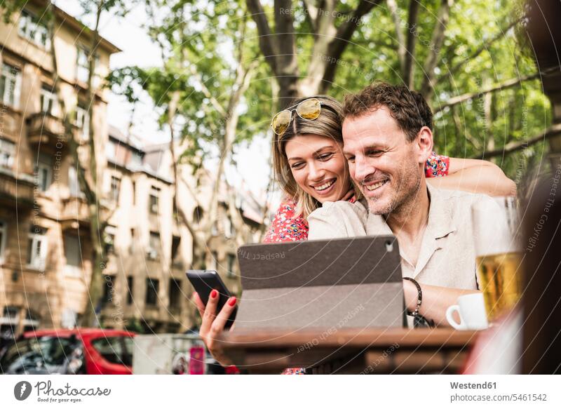 Smiling couple sharing tablet and cell phone at an outdoor cafe share smiling smile happiness happy mobile phone mobiles mobile phones Cellphone cell phones