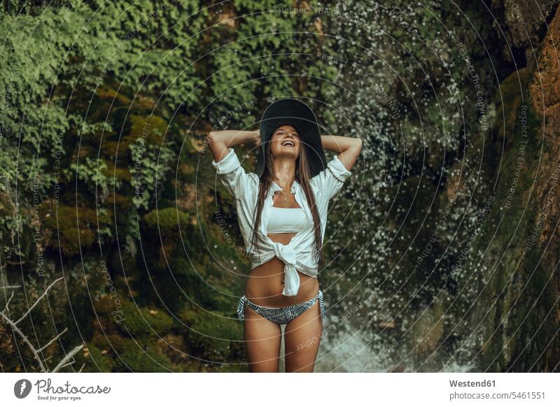 Happy young woman wearing a hat standing near a waterfall swim wear bikinis hats summer time summertime summery relax relaxing relaxation delight enjoyment