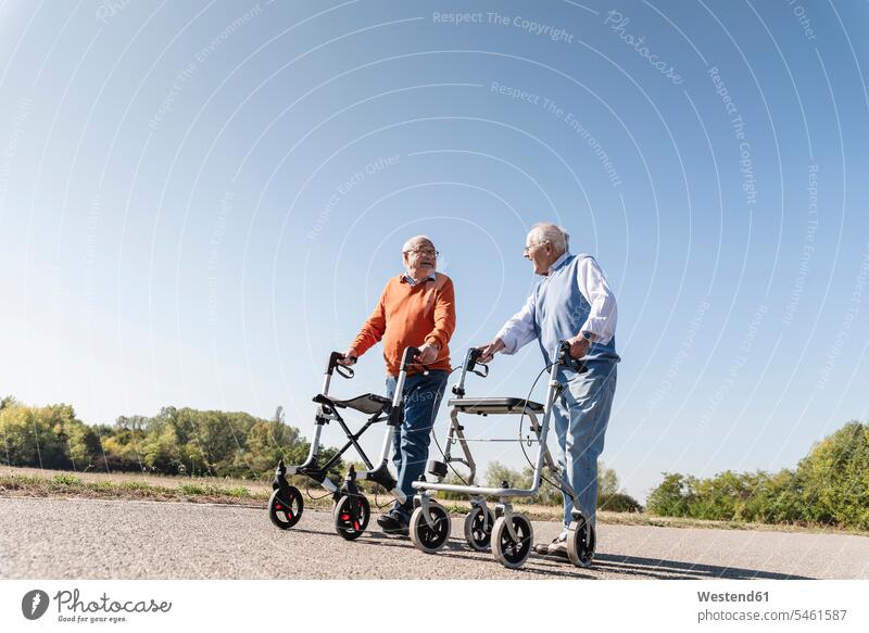 Two old friends walking on a country road, using wheeled walkers caucasian caucasian ethnicity caucasian appearance european Frail Frailty summer summer time