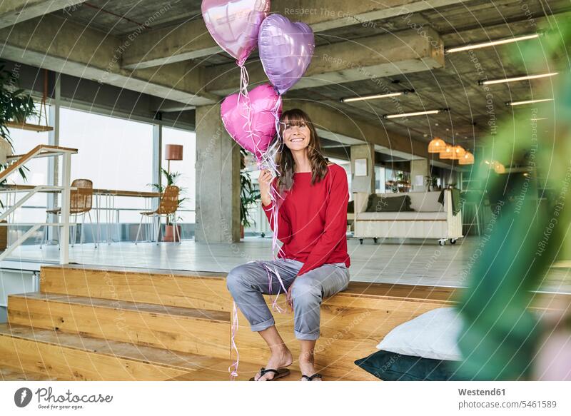 Businesswoman holding balloons while sitting on stairs in loft office color image colour image indoors indoor shot indoor shots interior interior view Interiors