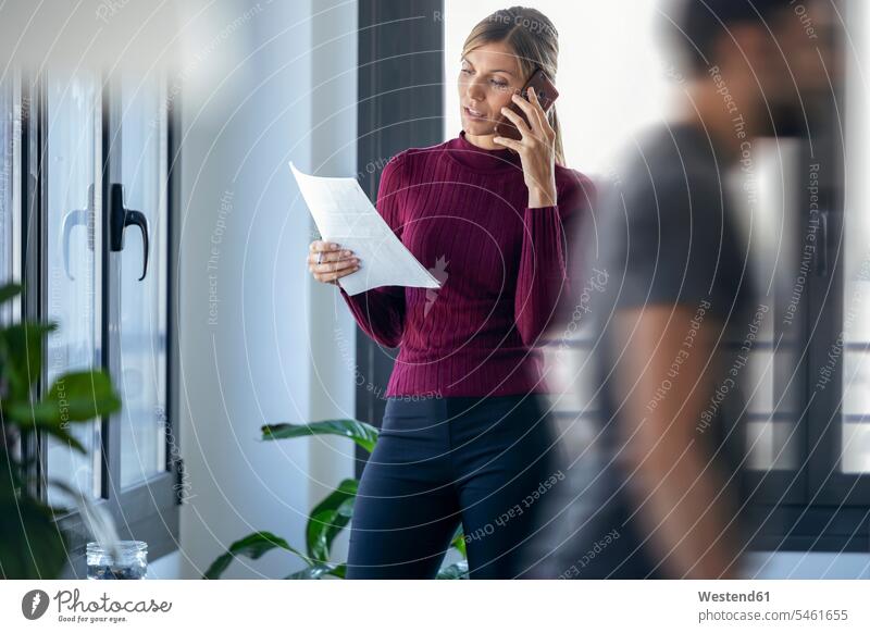 Businesswoman talking over smart phone while male coworker walking in office color image colour image Spain indoors indoor shot indoor shots interior