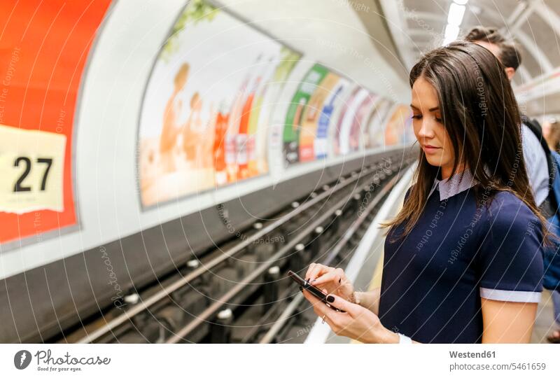 UK, London, young woman waiting at underground station platform looking at cell phone Smartphone iPhone Smartphones view seeing viewing Subway Platform females