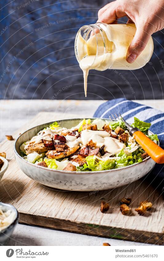 Hand of person pouring dressing over bowl of Caesar salad with romaine lettuce, Parmesan cheese, bacon, chicken breast and croutons close-up close up closeup