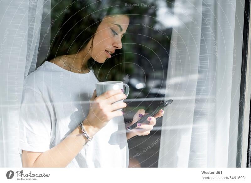 Young woman with cup of coffee and cell phone behind windowpane human human being human beings humans person persons celibate celibates singles solitary people