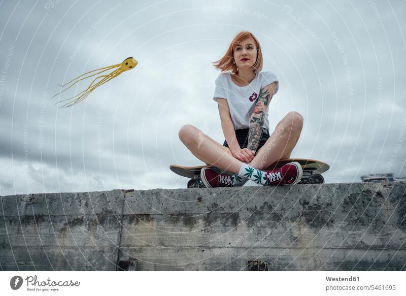 Young woman sitting on a concrete wall on carver skateboard with kite in background concrete walls Skate Board skateboards females women Seated Adults grown-ups