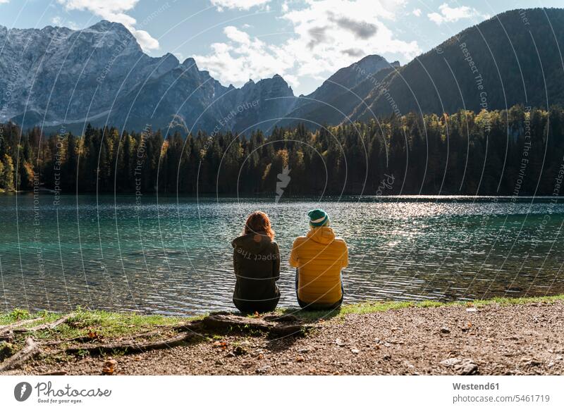 Rear view of couple sitting on lakeside at Laghi di Fusine, Friuli Venezia Giulia, Italy coat coats jackets Seated enjoy enjoyment indulgence indulging savoring