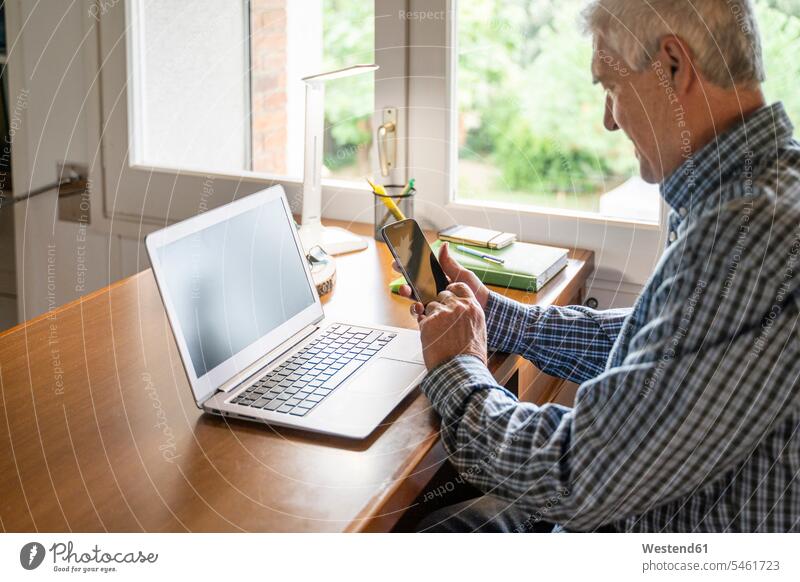 Senior man sitting in front of laptop using smartphone at home human human being human beings humans person persons caucasian appearance caucasian ethnicity