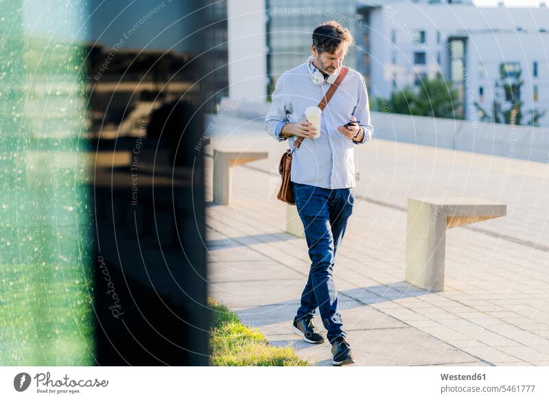 Mature man with takeaway coffee and headphones using cell phone in the city Coffee men males headset town cities towns mobile phone mobiles mobile phones
