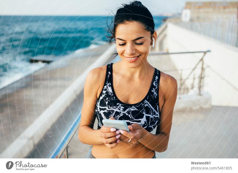 Woman using smartphone and in-ear during workout, sitting on a pier telecommunication phones telephone telephones cell phone cell phones Cellphone mobile