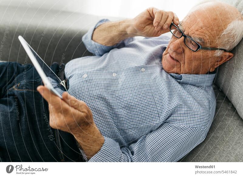 Senior man surfing the internet on a tablet while lying on the sofa couch settee sofas couches settees senior men senior man elder man elder men senior citizen