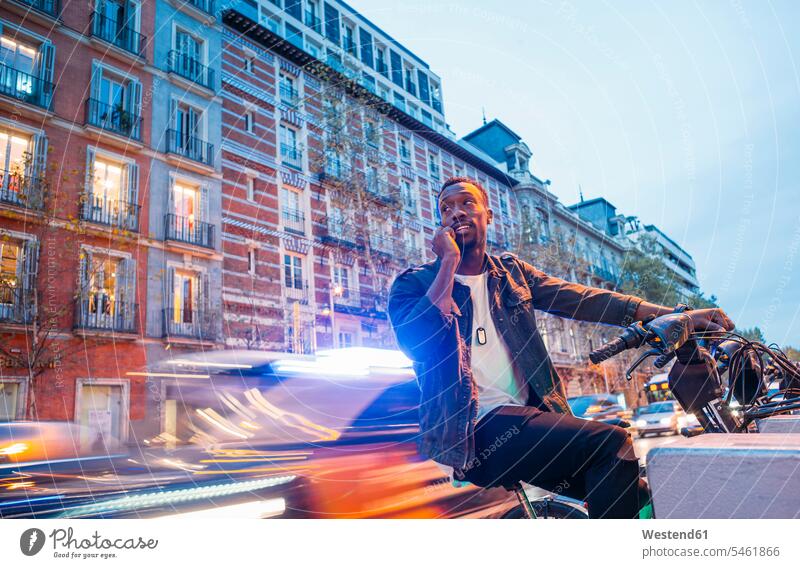 Smiling man talking over smart phone while sitting on bicycle in city at dusk color image colour image Spain leisure activity leisure activities free time