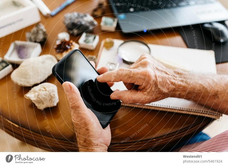 Hands of elderly retired man using smart phone while doing research on fossil and mineral at home color image colour image indoors indoor shot indoor shots