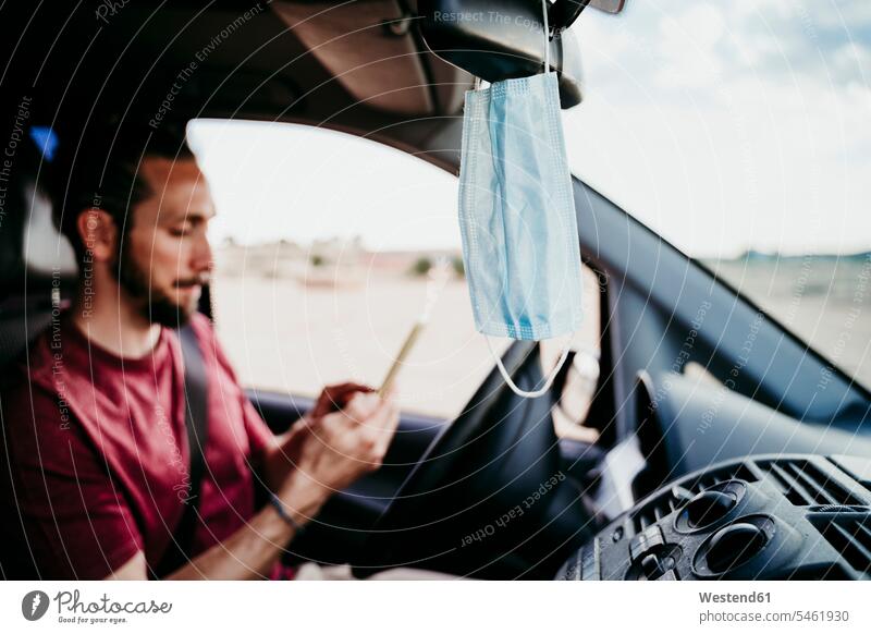 Close-up of face mask hanging on rear-view mirror while man using phone in car color image colour image Vehicle Interior day daylight shot daylight shots