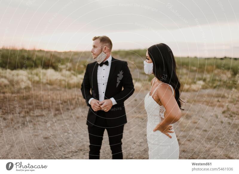 Bride with groom wearing protective face mask while looking away in field during COVID-19 color image colour image outdoors location shots outdoor shot