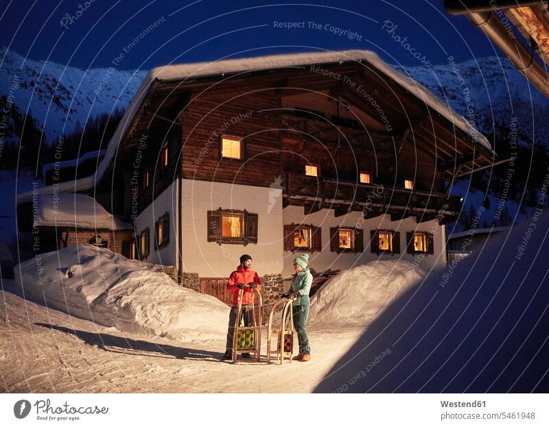 Couple with sledges in snow-covered landscape with rustic house at night landscapes scenery terrain houses by night nite night photography couple twosomes