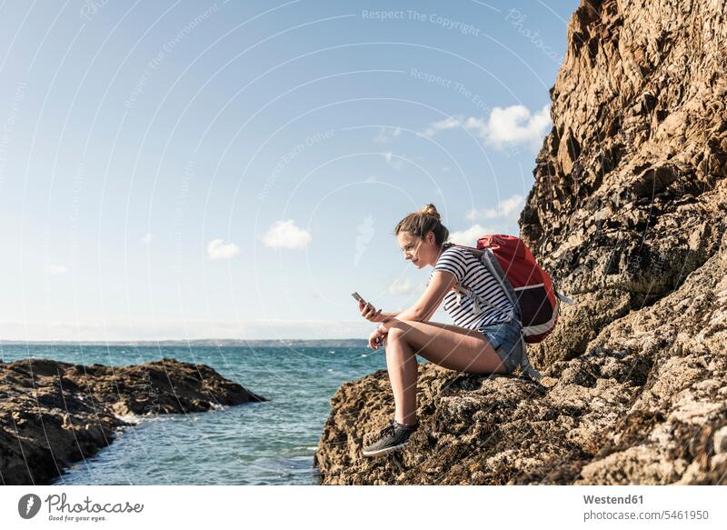 Young woman sitting on a rocky beach, using smartphone beach hiking rocks Seated use beaches Smartphone iPhone Smartphones young women young woman hike