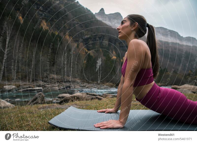 Active young woman exercising Cobra pose on yoga mat near river and mountain color image colour image outdoors location shots outdoor shot outdoor shots Huesca