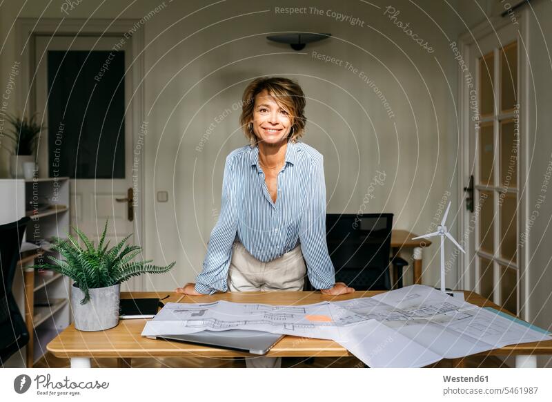 Smiling female architect leaning on desk while standing in office color image colour image indoors indoor shot indoor shots interior interior view Interiors day