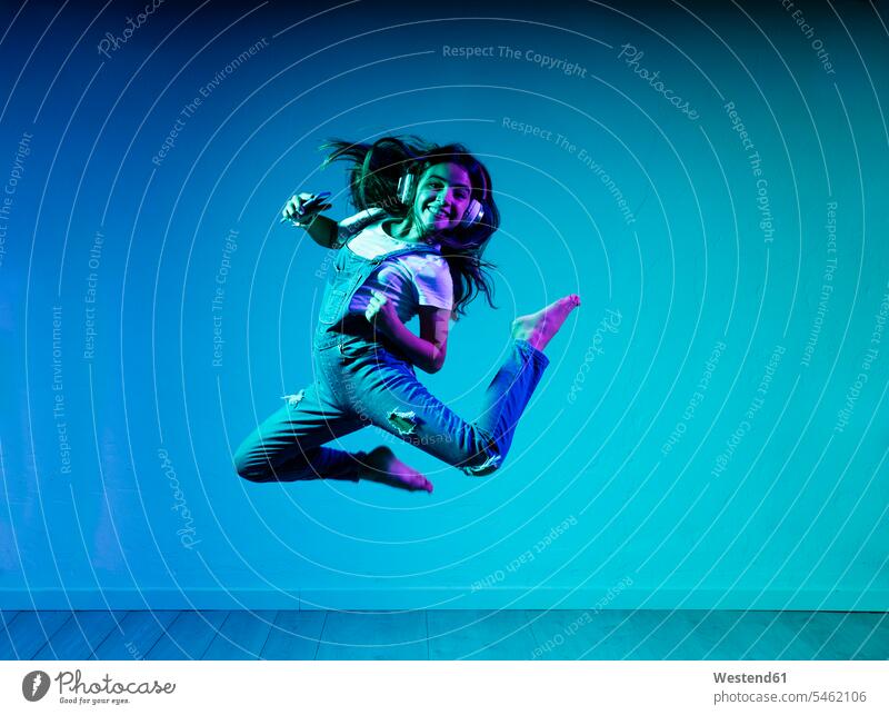 Cheerful girl jumping while listening music through headphones against blue wall color image colour image indoors indoor shot indoor shots interior