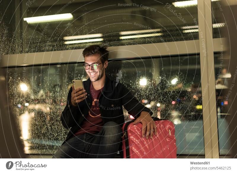 Smiling young man with suitcase sitting at rainy window using smartphone human human being human beings humans person persons caucasian appearance