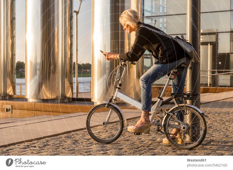Senior woman with city bike using cell phone at the riverside at sunset bicycle bikes bicycles females women riverbank sunsets sundown mobile phone mobiles