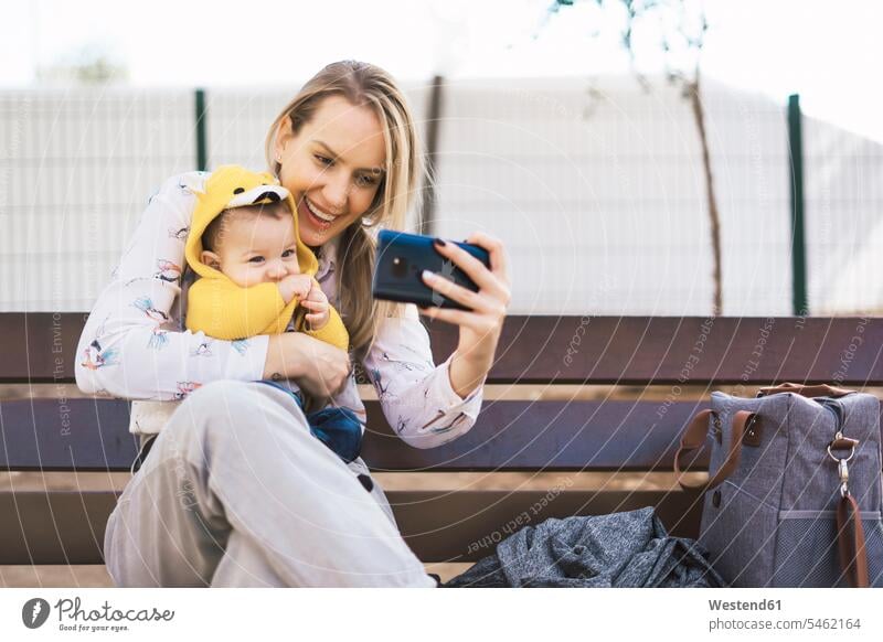 Happy mother resting with baby boy on a park bench taking a selfie bags coat coats jackets benches park benches telecommunication phones telephone telephones