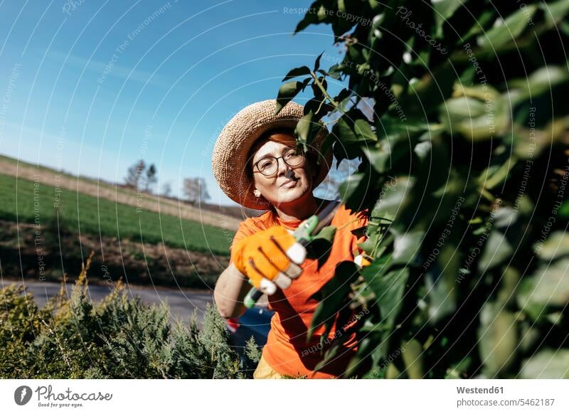 Portrait of mature woman with straw hat trimming facade greenery Eye Glasses Eyeglasses specs spectacles cut stand free time leisure time Distinct individual