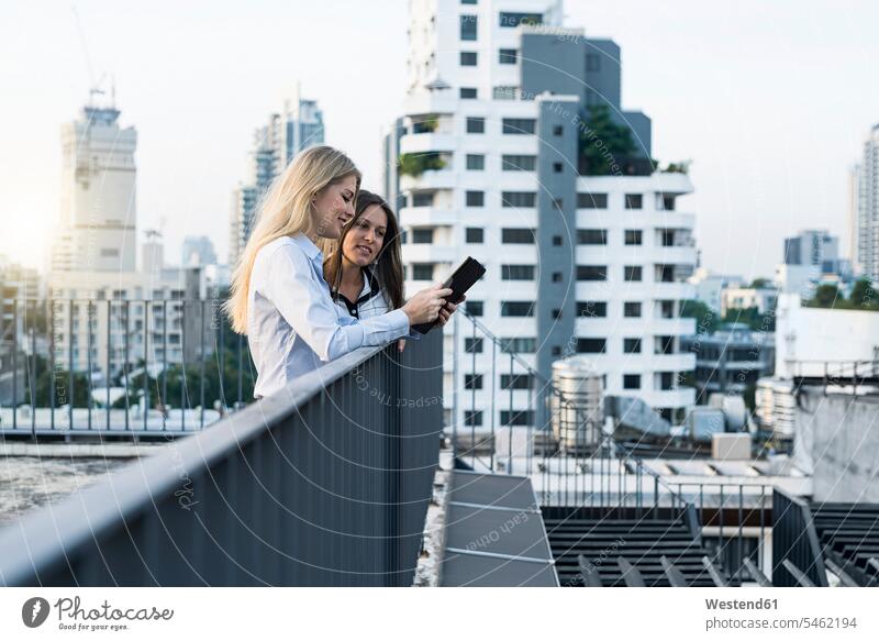 Two businesswomen talking on city rooftop, using digital tablet business people businesspeople businesswoman business woman business women smiling smile