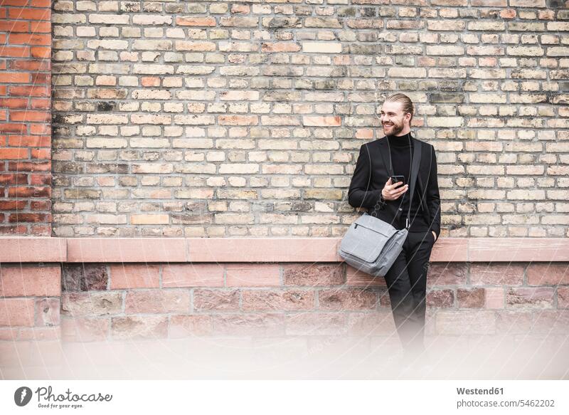 Smiling fashionable young man with cell phone leaning against brick wall looking sideways standing smiling smile brick walls men males Adults grown-ups grownups