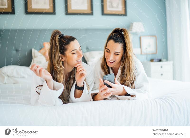 Smiling twin sisters using smart phone while lying on bed in hotel color image colour image Spain leisure activity leisure activities free time leisure time