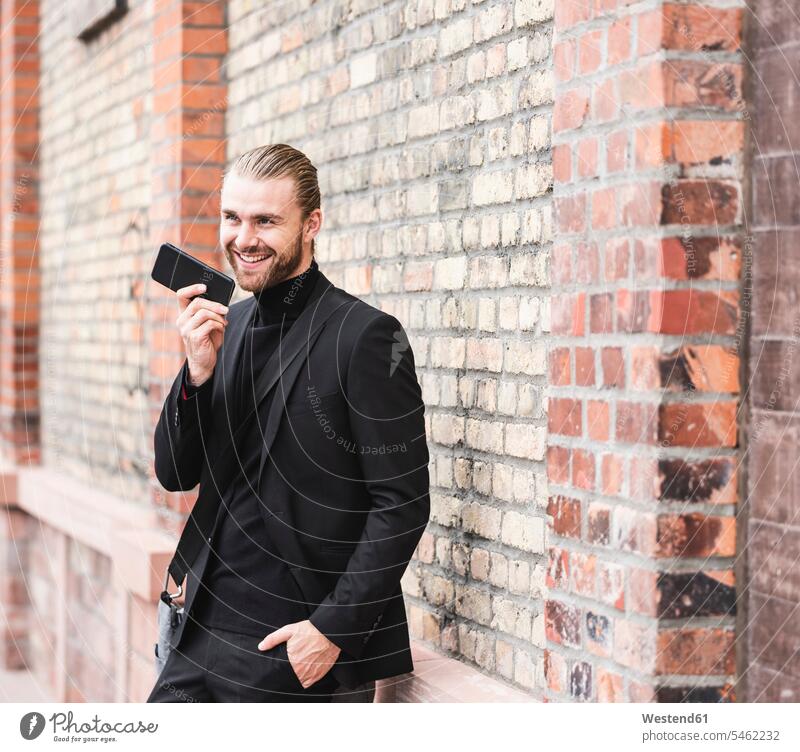 Smiling fashionable young man standing at brick wall using cell phone smiling smile brick walls portrait portraits mobile phone mobiles mobile phones Cellphone