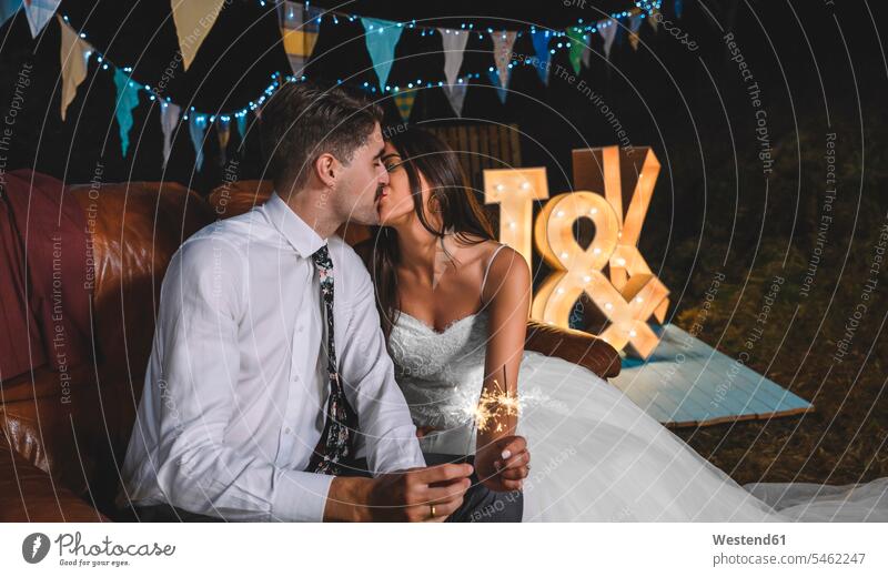 Romantic wedding couple sitting on sofa kissing while holding sparklers in their hands on a night party outdoors Seated bridal couple bridal couples romantic